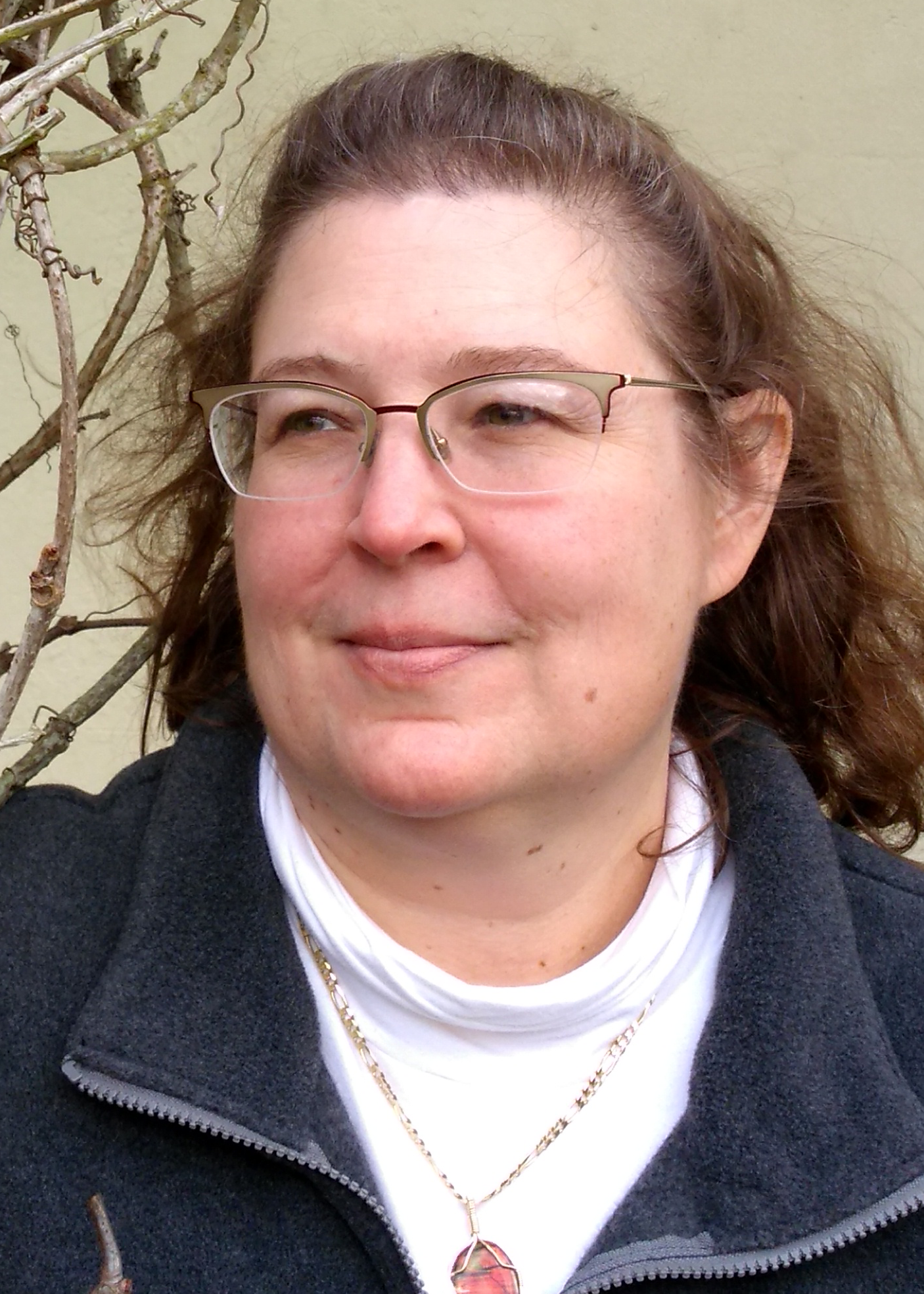 A woman with brown hair and glasses is wearing a white turtleneck, a dark zip-up jacket, and a necklace with a pink pendant. She is outdoors, standing near some bare branches and looking slightly to the side.