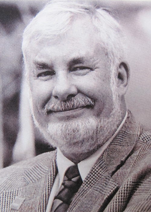 A man with white hair and a beard is smiling. He is wearing a checkered blazer, a dress shirt, and a tie. The image is in black and white.