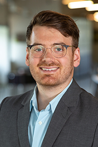 A person with short brown hair, glasses, and a beard is wearing a gray suit jacket over a light blue shirt. They are smiling, and the background is a blurred indoor setting with lights.
